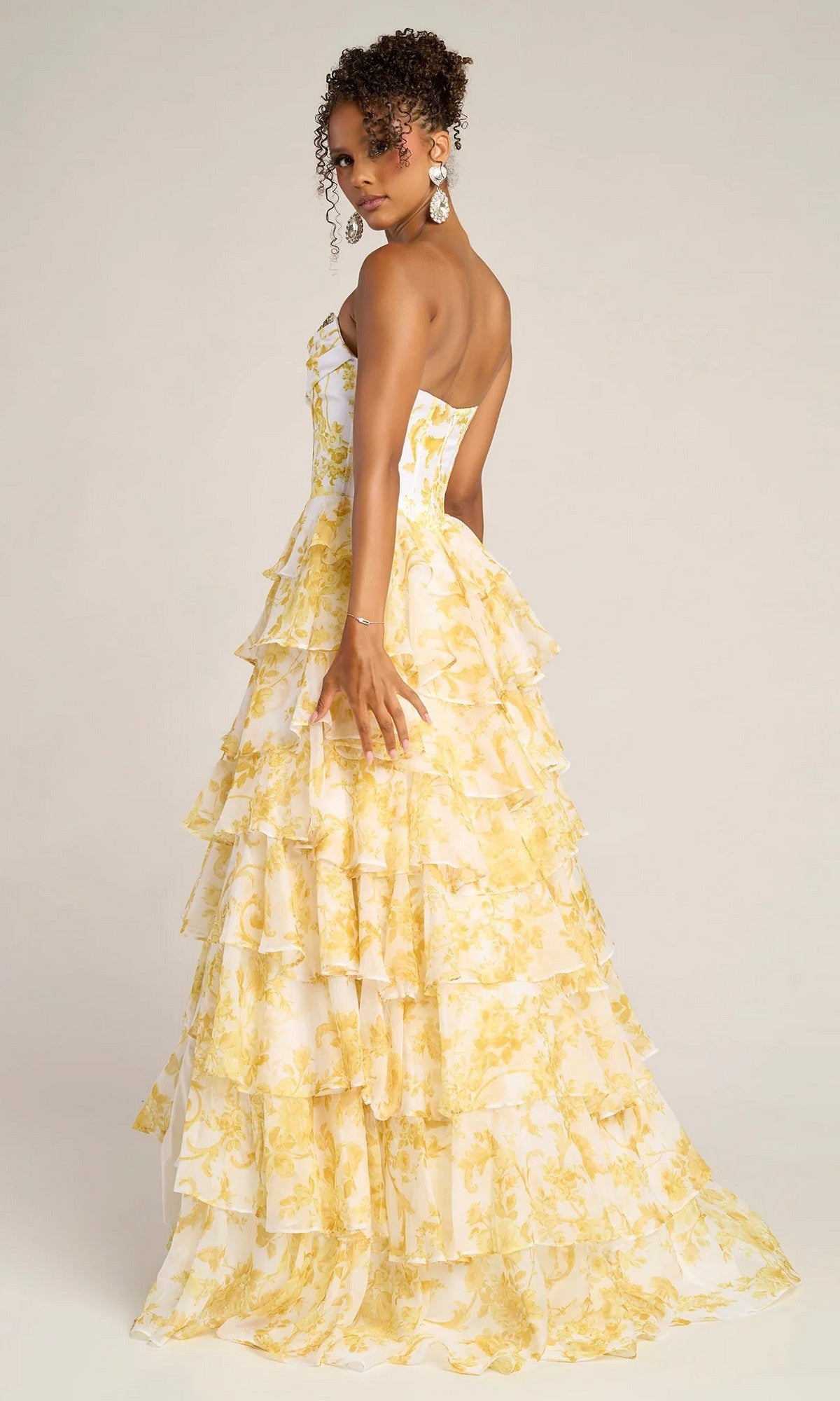 A woman poses in the Portia and Scarlett PS26210 long prom dress, a strapless ball gown featuring yellow floral patterns and tiered ruffles, looking over her shoulder against a plain background.